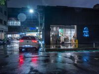 a small car parked at the street corner in front of a store front in rain at night