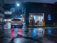 a small car parked at the street corner in front of a store front in rain at night