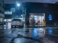 a small car parked at the street corner in front of a store front in rain at night