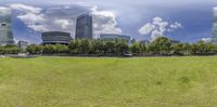 a green grassy field that is next to tall buildings and trees in front of it