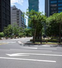 a road with a bus stop sign and green trees near tall buildings in the distance