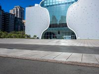 a building made of large white shaped glass and metal on a street near buildings under blue skies