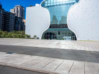 a building made of large white shaped glass and metal on a street near buildings under blue skies