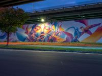 a very colorful painting on the side of a bridge in town by some grass and trees