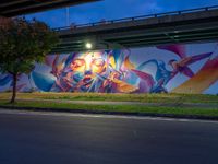 a very colorful painting on the side of a bridge in town by some grass and trees