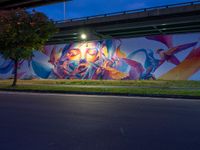 a very colorful painting on the side of a bridge in town by some grass and trees