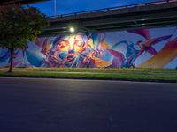 a very colorful painting on the side of a bridge in town by some grass and trees