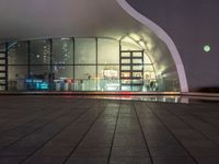 a sidewalk outside of a building at night that has several windows and lights on the glass walls