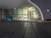 a sidewalk outside of a building at night that has several windows and lights on the glass walls