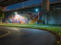 a wall with a colorful mural on it next to an empty road near grass and trees