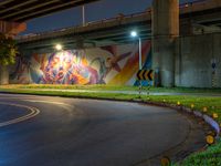 a wall with a colorful mural on it next to an empty road near grass and trees