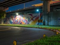 a wall with a colorful mural on it next to an empty road near grass and trees