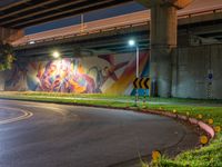 a wall with a colorful mural on it next to an empty road near grass and trees