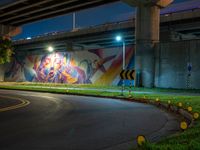 a wall with a colorful mural on it next to an empty road near grass and trees