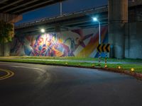 a wall with a colorful mural on it next to an empty road near grass and trees