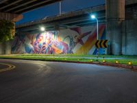 a wall with a colorful mural on it next to an empty road near grass and trees