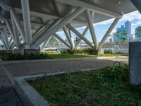 a bench underneath a bridge sitting in a green park area surrounded by large metal columns