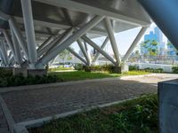 a bench underneath a bridge sitting in a green park area surrounded by large metal columns