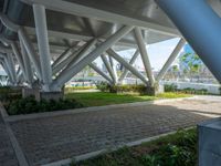 a bench underneath a bridge sitting in a green park area surrounded by large metal columns