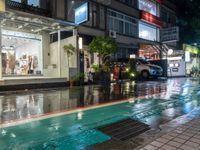 a wet street with a storefront on the corner, at night and it's rain still coming