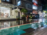 a wet street with a storefront on the corner, at night and it's rain still coming