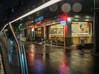 a wet street with a car on the road near store fronts at night and a neon sign for club nus