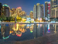 a rainy city street with tall buildings reflecting in it's water at nighttime