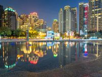 a rainy city street with tall buildings reflecting in it's water at nighttime