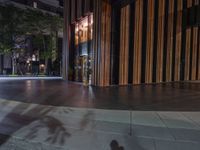 an empty sidewalk and a tall building by night with light reflecting off the windows of it