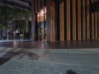 an empty sidewalk and a tall building by night with light reflecting off the windows of it