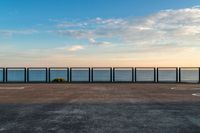 the fence and the road are in front of the water with some clouds in the sky