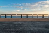 the fence and the road are in front of the water with some clouds in the sky