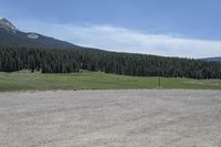 a dirt field and road near pine trees and mountain tops against blue sky with wispy clouds