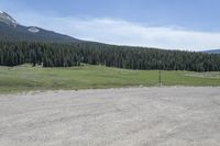 a dirt field and road near pine trees and mountain tops against blue sky with wispy clouds