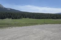 a dirt field and road near pine trees and mountain tops against blue sky with wispy clouds