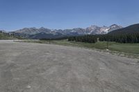 a dirt field and road near pine trees and mountain tops against blue sky with wispy clouds