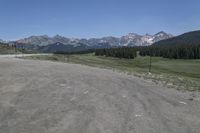 a dirt field and road near pine trees and mountain tops against blue sky with wispy clouds