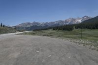 a dirt field and road near pine trees and mountain tops against blue sky with wispy clouds