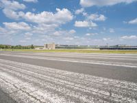 an empty runway surrounded by trees and blue skies and clouds with small buildings along it