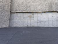 man standing on concrete holding his skate board in one hand and leaning against the wall