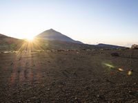 two people standing in a dirt field with a herd of sheep and sun shining over the mountain top