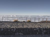 the person is posing on a rocky ledge overlooking the clouds and mountains near a fence