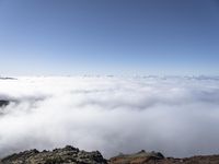 a person looking out over a layer of clouds as they climb a mountain peak, with rocks and boulders below