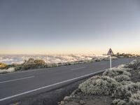 a roadway on a mountain with clouds and trees in the background to a clear, blue sky