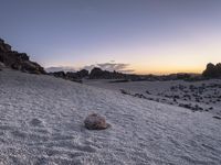 a rock on the sandy ground in front of some rocks at sunset with a sky background