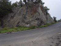 a motorcycle is going down an empty road by the rock wall and forest trees are in front of it