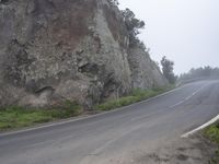 a motorcycle is going down an empty road by the rock wall and forest trees are in front of it