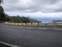 a concrete barrier along a mountain side with sea in the distance and trees on the right