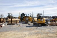 three tractors and a bulldozer are parked in front of a building site where piles of lumber sits