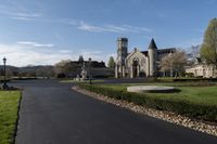 the driveway has gravel and grass surrounding it and a large stone church building with a steeple on top